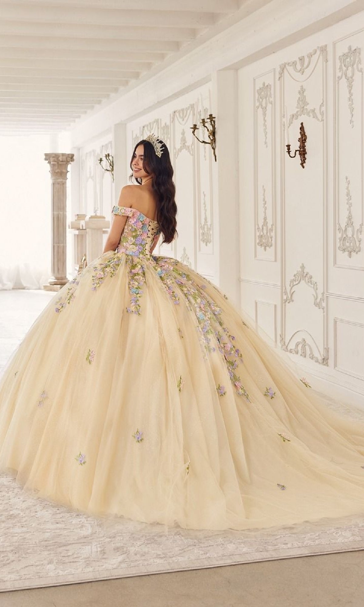 A young woman in the Quinceanera Dress 15724 by Ladivine, an off-the-shoulder pastel floral applique ball gown, stands in an elegant, ornate room, looking over her shoulder and smiling.
